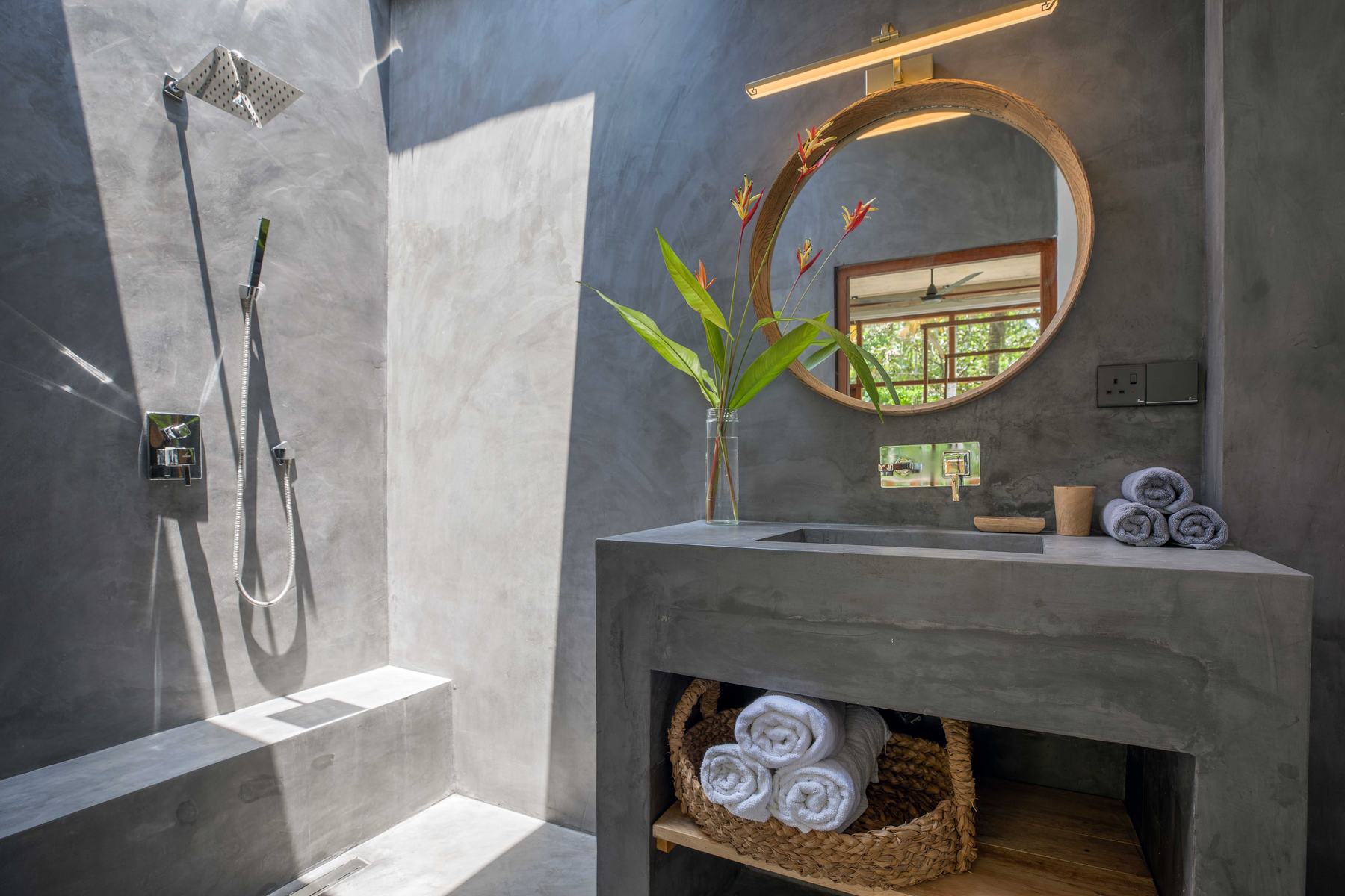 Master bathroom in grey concrete, with sunlight falling into the shower from the open ceiling above, a round wooden mirror and rolled towels