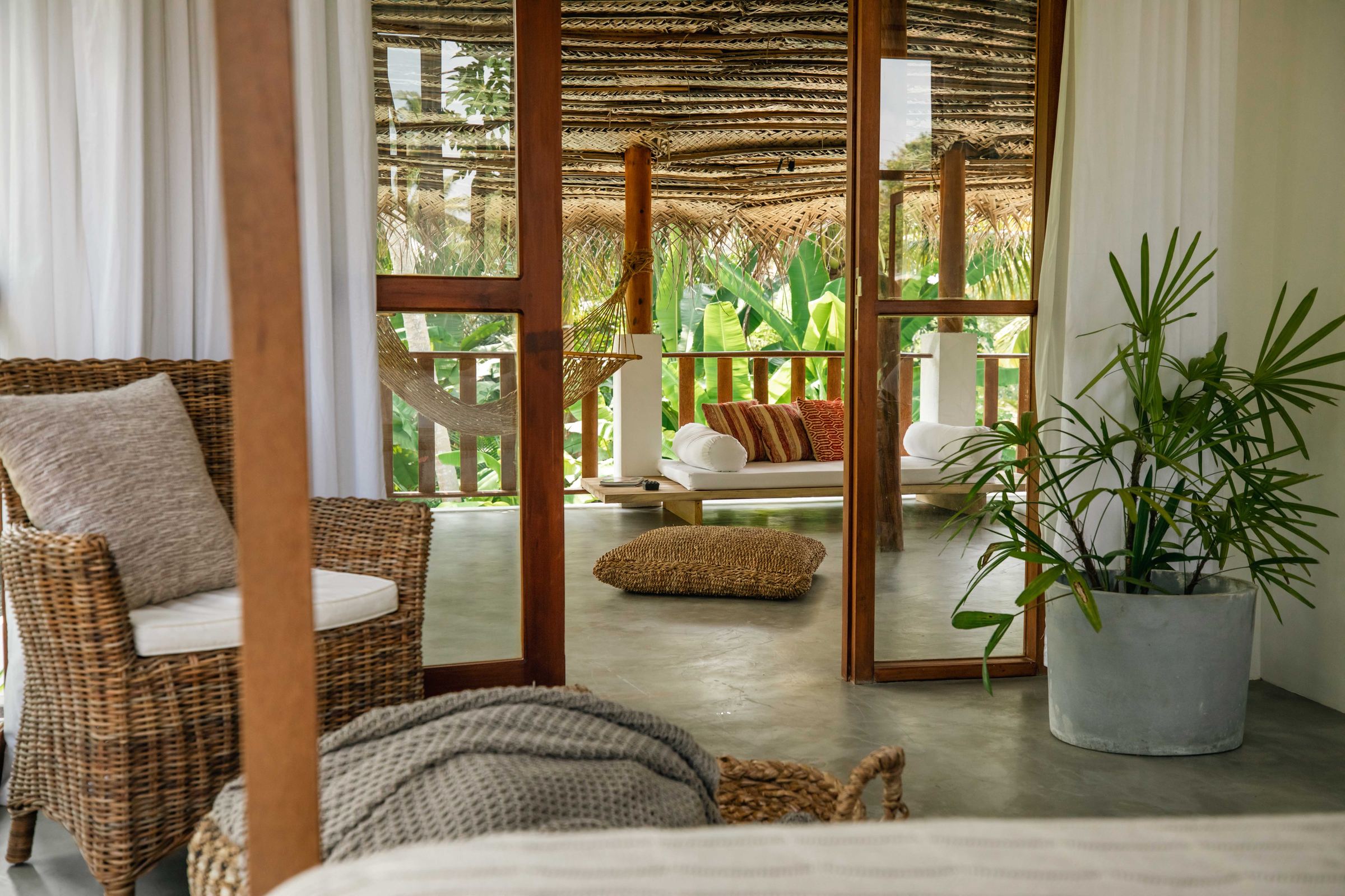 Looking out from a bedroom through open teak doors to the thatched veranda, with a hammock, daybed and banana palms beyond