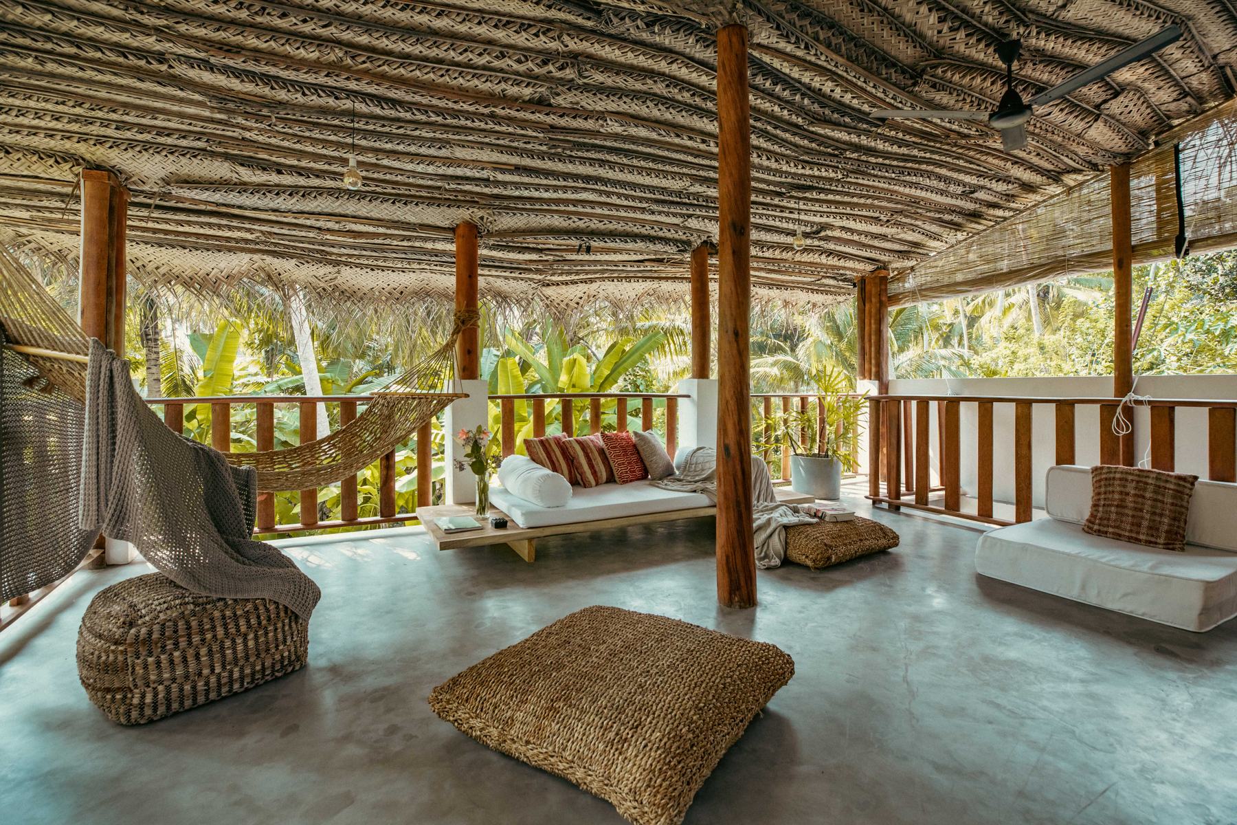Upstairs veranda under a woven thatch roof with daybeds, floor cushions and a hammock, open to the palms
